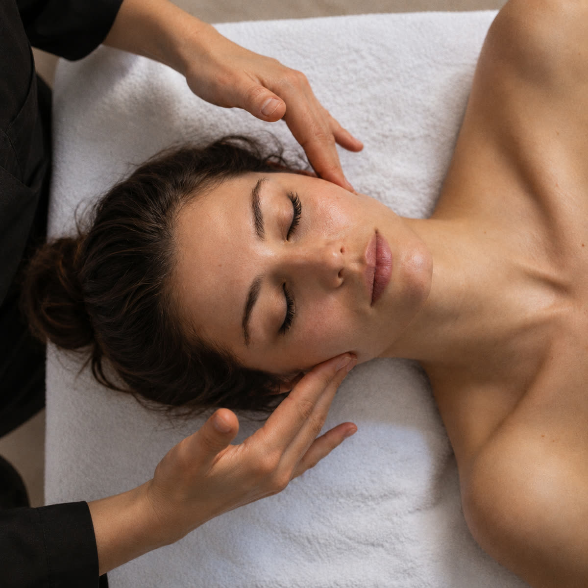 Vue plongeante d’un massage du visage, femme allongée yeux clos sur une serviette blanche