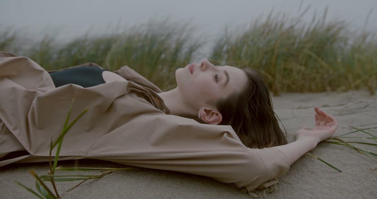Jeune femme en chemise beige allongée sur le sable parmi les oyats, sous un ciel gris