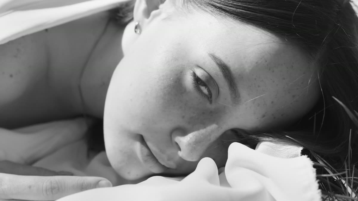 Gros plan noir et blanc d’un visage de femme aux taches de rousseur, allongée sur un drap