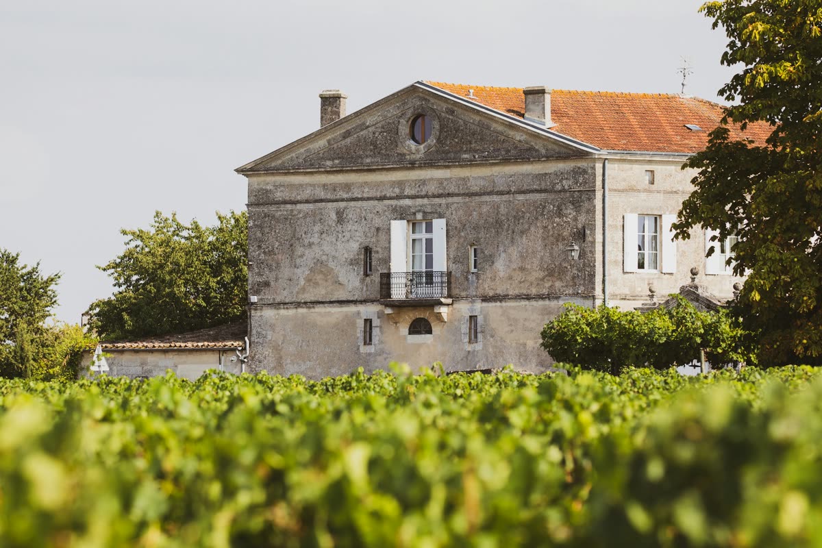 Façade en pierre d’une demeure viticole à volets blancs et toit de tuiles, derrière des vignes