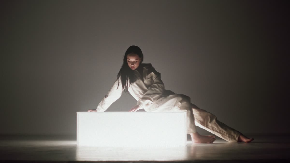 Femme en combinaison blanche penchée sur un bloc lumineux dans un studio plongé dans l’ombre