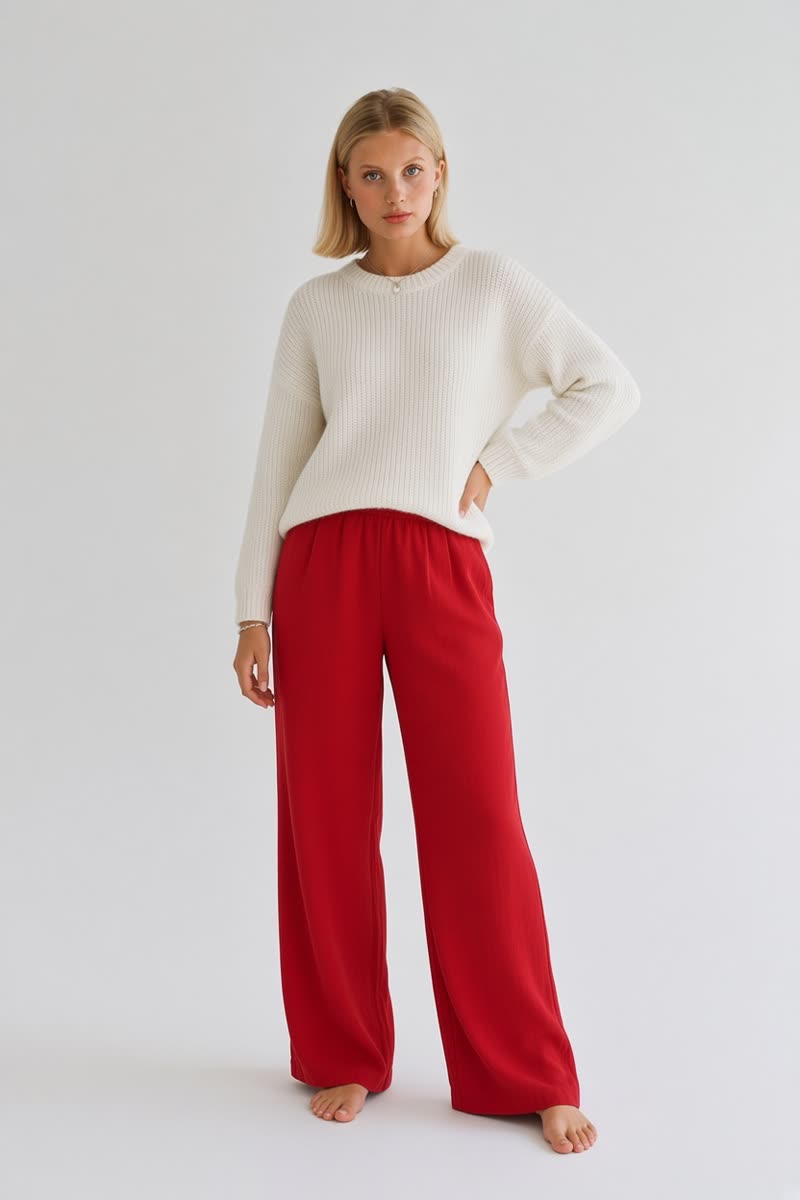 Femme blonde debout en pull blanc et pantalon large rouge, main sur la hanche, fond clair